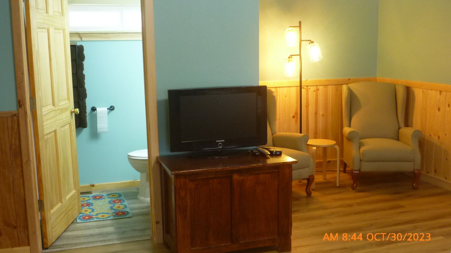 Une pièce avec un plancher en bois, un téléviseur posé sur un meuble en bois, une chaise, une petite table et une lampe.