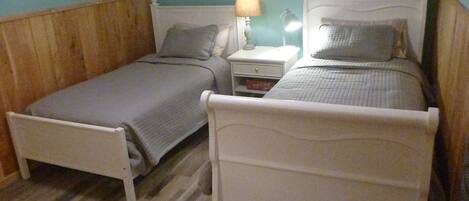 Two single beds with wooden headboards and white frames, a nightstand with a lamp, and a small drawer unit.