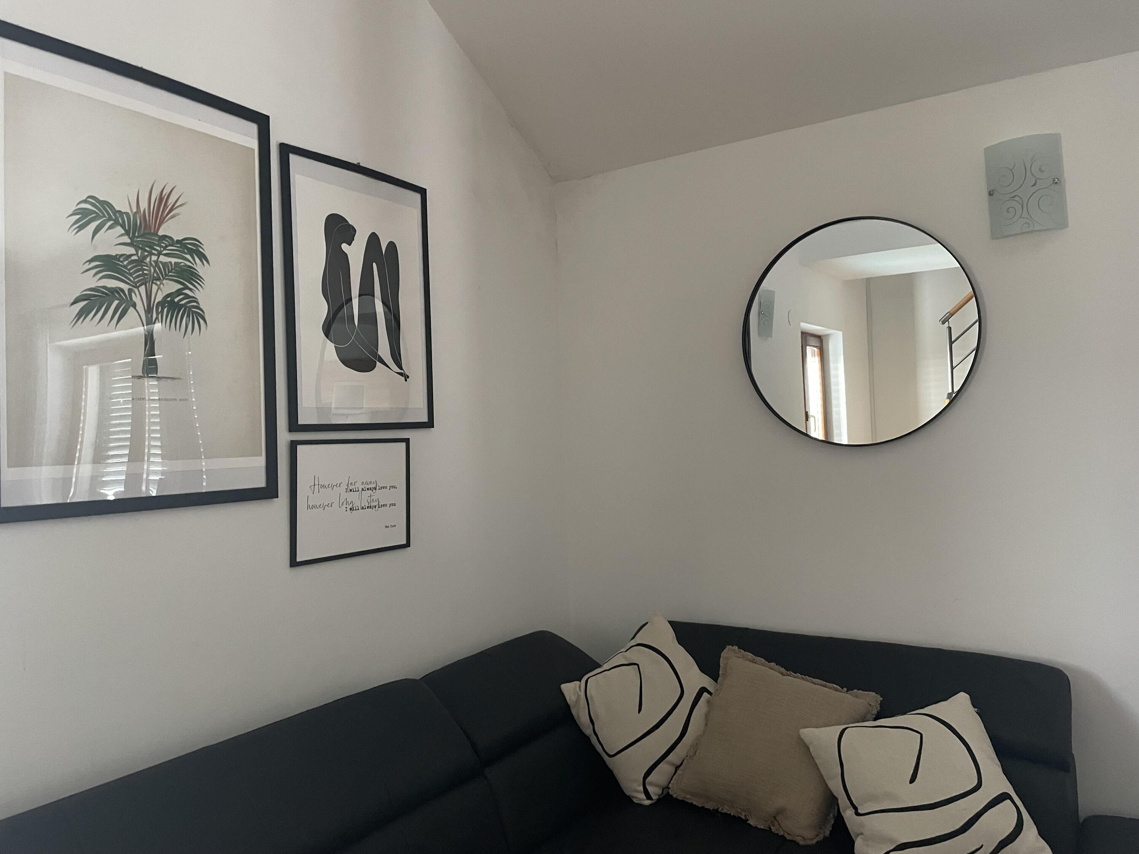 Uma sala de estar com um sofá preto, três obras de arte emolduradas e um espelho redondo na parede.