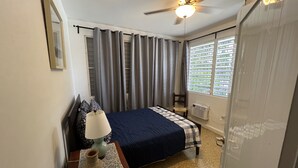 Une chambre à coucher comprenant un lit, une table de chevet, une lampe, un ventilateur de plafond et une fenêtre avec des stores.