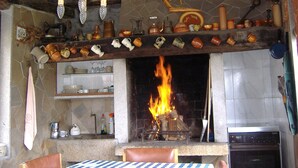 Una cocina rústica con chimenea, estantes con utensilios de cocina y un comedor con mantel a cuadros.