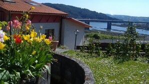 Una casa con techo rojo, un jardín lleno de flores y un río con un puente al fondo.