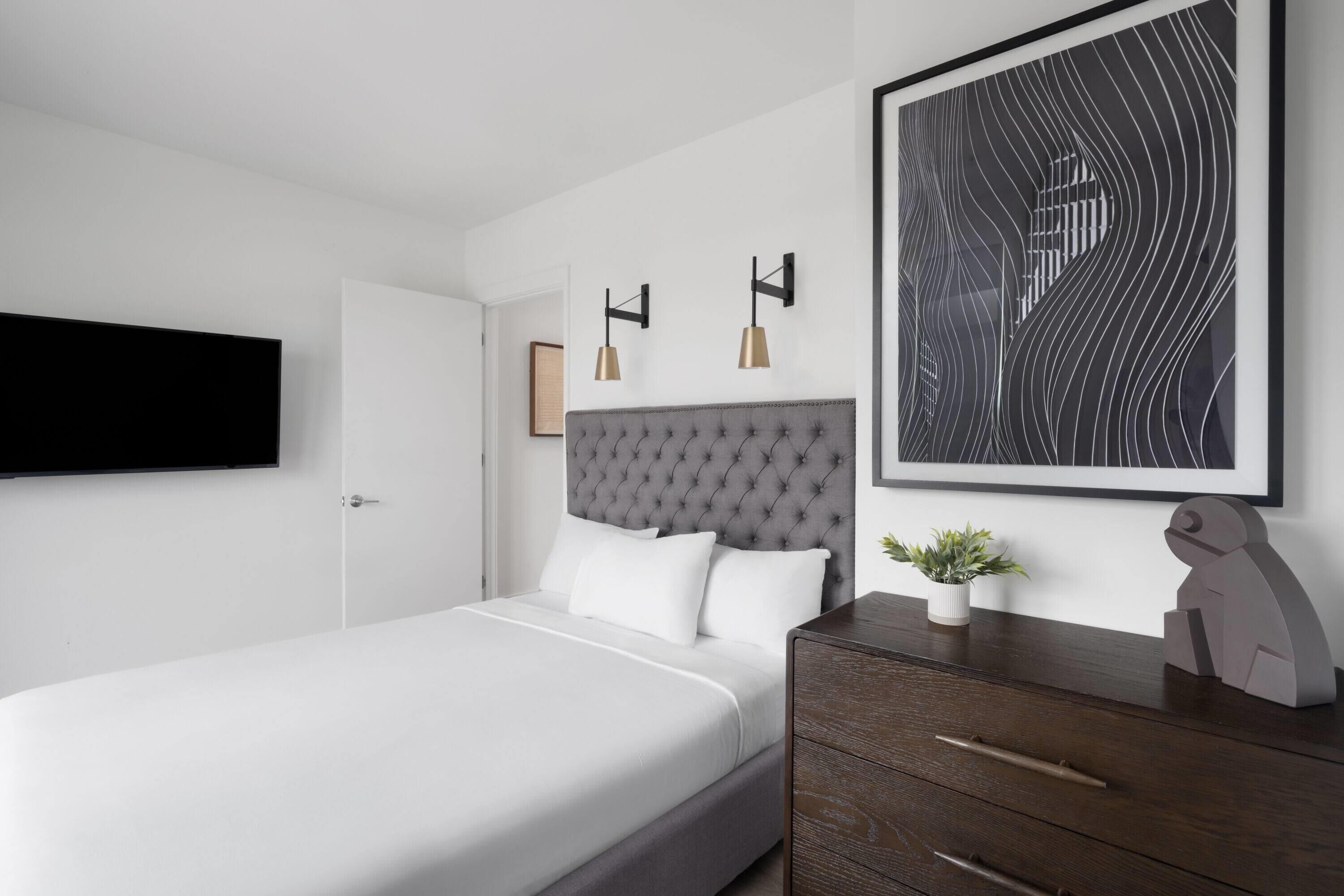 A modern bedroom with a large bed, a dark wooden dresser, a framed abstract painting, and a mounted TV.