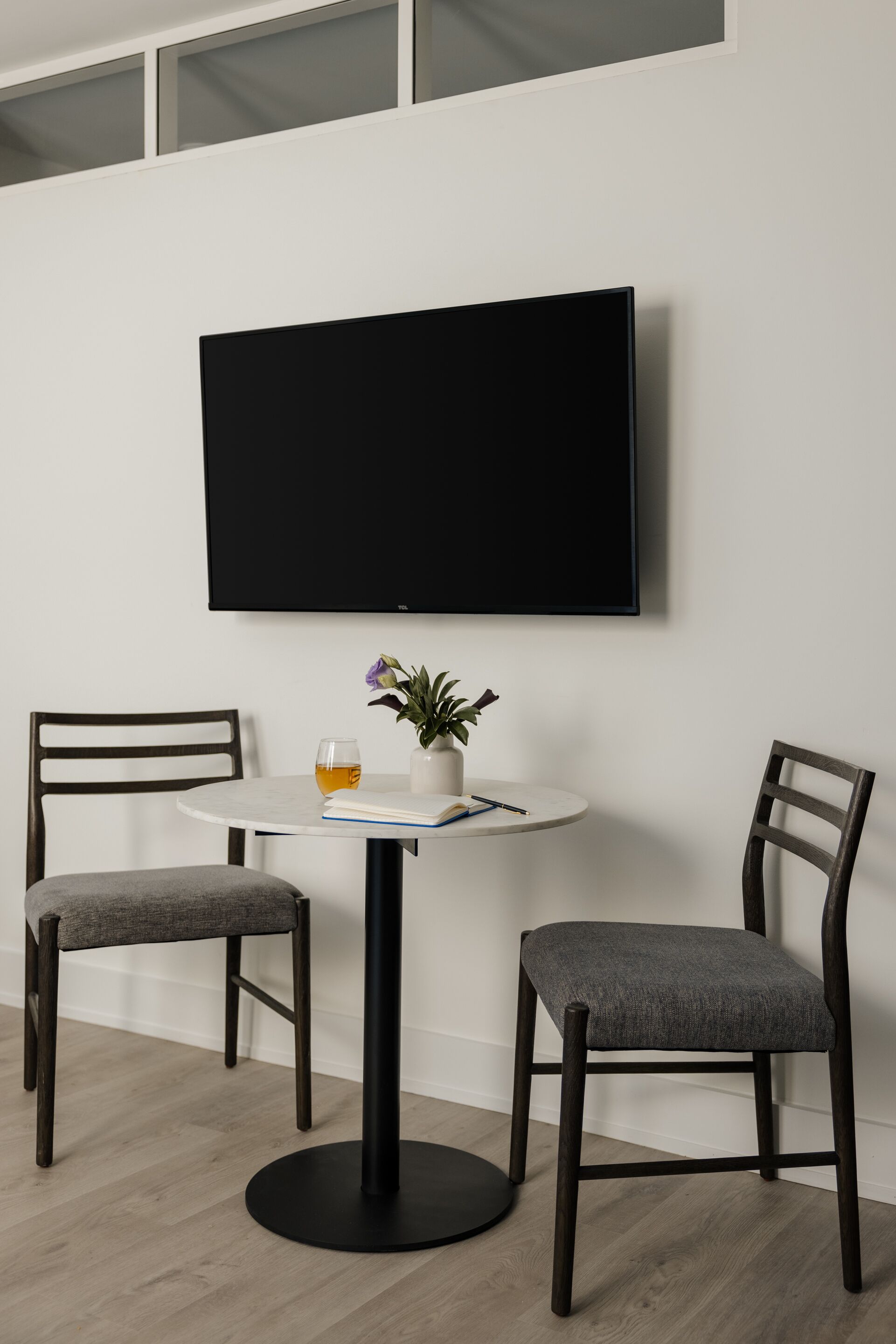 A modern room with a round table, two chairs, a flat-screen TV mounted on the wall, a small potted plant, and a glass of tea.