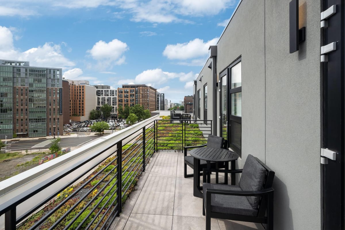 A balcony with a view of city buildings, a small table with chairs, and a railing with plants.