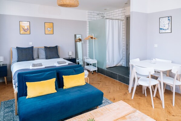 A modern bedroom with a large bed, a bedside table, a mirror, a shower area, and a dining table with chairs.