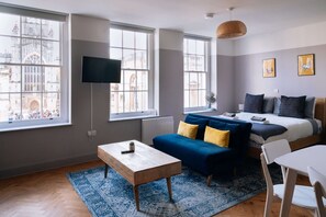 A modern hotel room with a large window, a bed, a sofa, a table, and a TV.