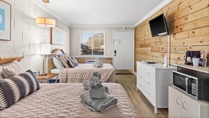 A hotel room with two beds, a wooden wall, a TV, a desk with a microwave, and a window with a view of buildings.