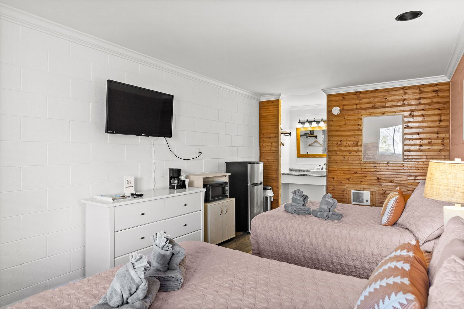 Hotellihuone, jossa on kaksi sänkyä, televisio, pieni keittiönurkkaus ja yksi puinen seinä, joka toimii koristeellisena elementtinä.