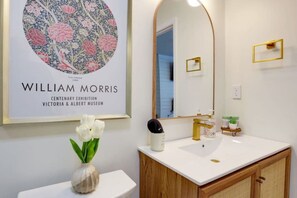 Een badkamer met een ingelijste afbeelding, een vaas met witte bloemen, een wastafel met gouden details en een spiegel.