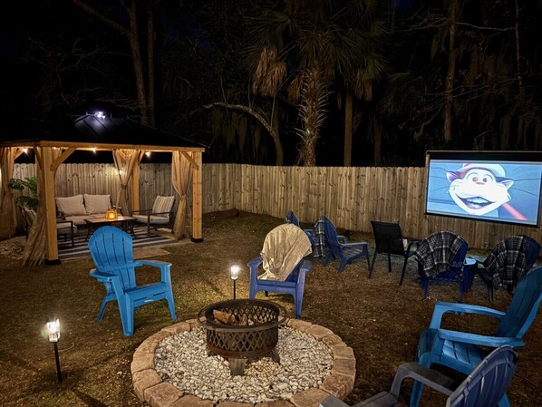 An outdoor patio area with a fire pit, blue chairs, a covered seating area, and a screen displaying a cat.