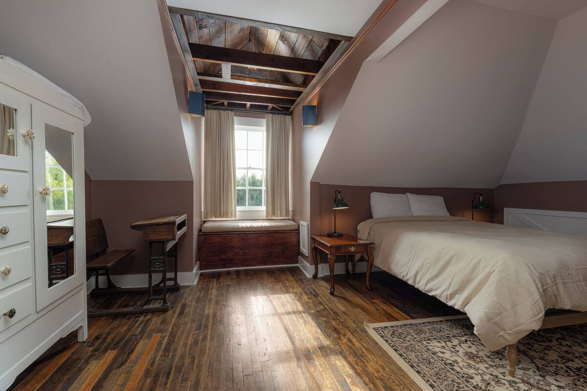 Ein Schlafzimmer mit schrägen Wänden, Holzboden, einem Bett, einem Nachttisch, einer Kommode, einer Bank und einem Fenster mit Vorhängen.