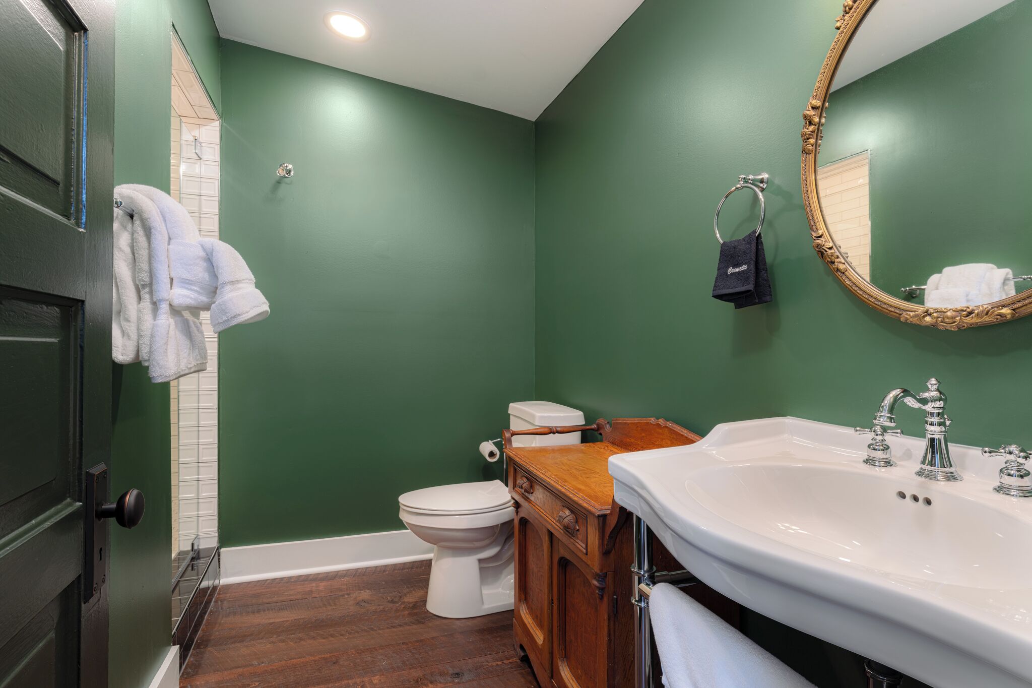 Un baño con pared verde, inodoro blanco, tocador de madera y un espejo grande.