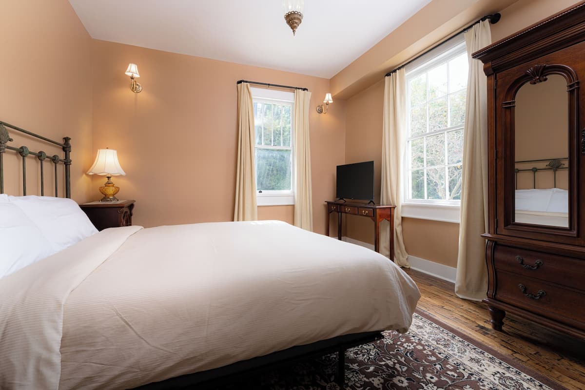 Ein Schlafzimmer mit einem großen Bett, einem Schminktisch, einem Fernseher und zwei mit Vorhängen versehenen Fenstern.