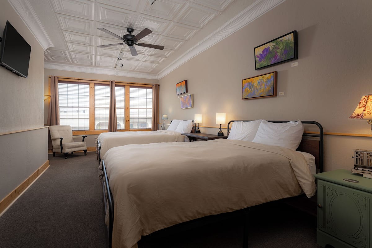 Ein Hotelzimmer mit zwei Betten, einem Deckenventilator, Kunstwerken an den Wänden und einem fenster mit Vorhängen.