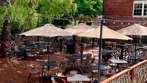 Eine Terrasse im Freien mit Tischen, Stühlen und Sonnenschirmen, im Hintergrund ein Backsteinhaus.