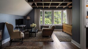 Ein Zimmer mit Holzboden, einem großen Fenster mit Vorhängen, einem Sofa, einem Sessel, einem kleinen Tisch mit einer Vase und einem Teppich.