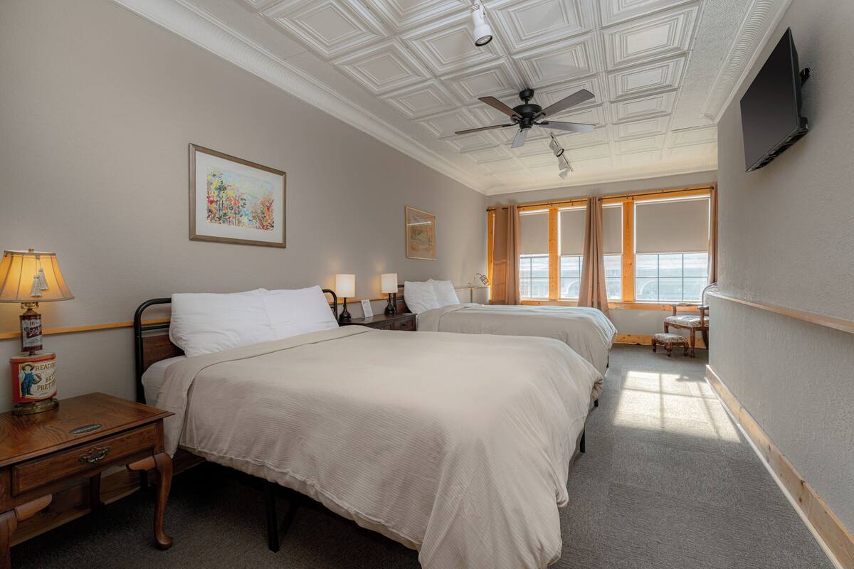 Ein Hotelzimmer mit zwei Betten, einem Deckenventilator, einem an der Wand befestigten Fernseher und einem Fenster mit Vorhängen.