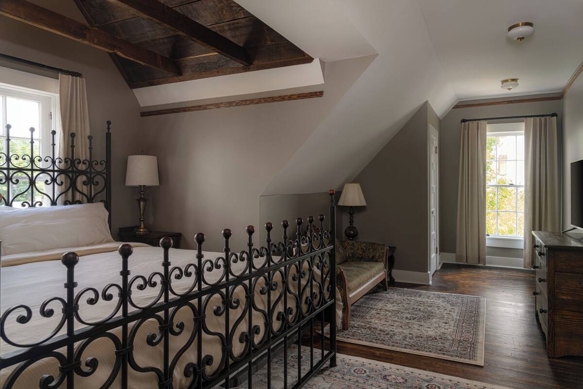 Ein Schlafzimmer mit einem Bett, einem Nachttisch, einer Lampe, einer Bank, einer Kommode und einem Fenster mit Vorhängen.