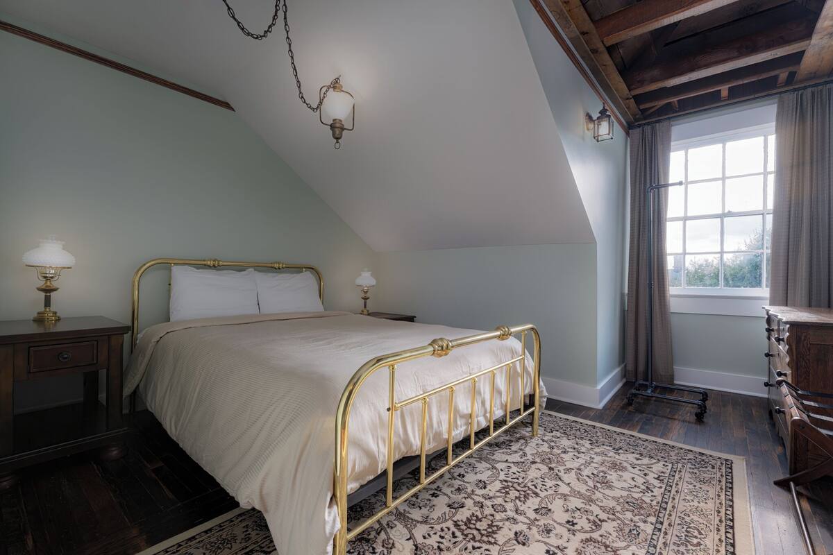 Ein Schlafzimmer mit schräg versiegelter Decke, einem Bett mit goldfarbener Rahmung, einem Nachttisch mit Lampe und einer Kommode.