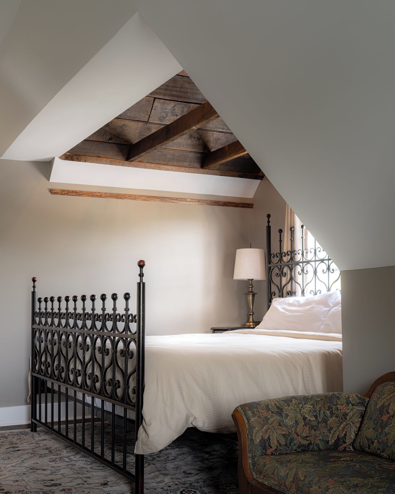 A bedroom with a sloped ceiling, a bed with a white bedspread, a metal headboard, a bedside lamp, a floral-patterned armchair, and a patterned rug.