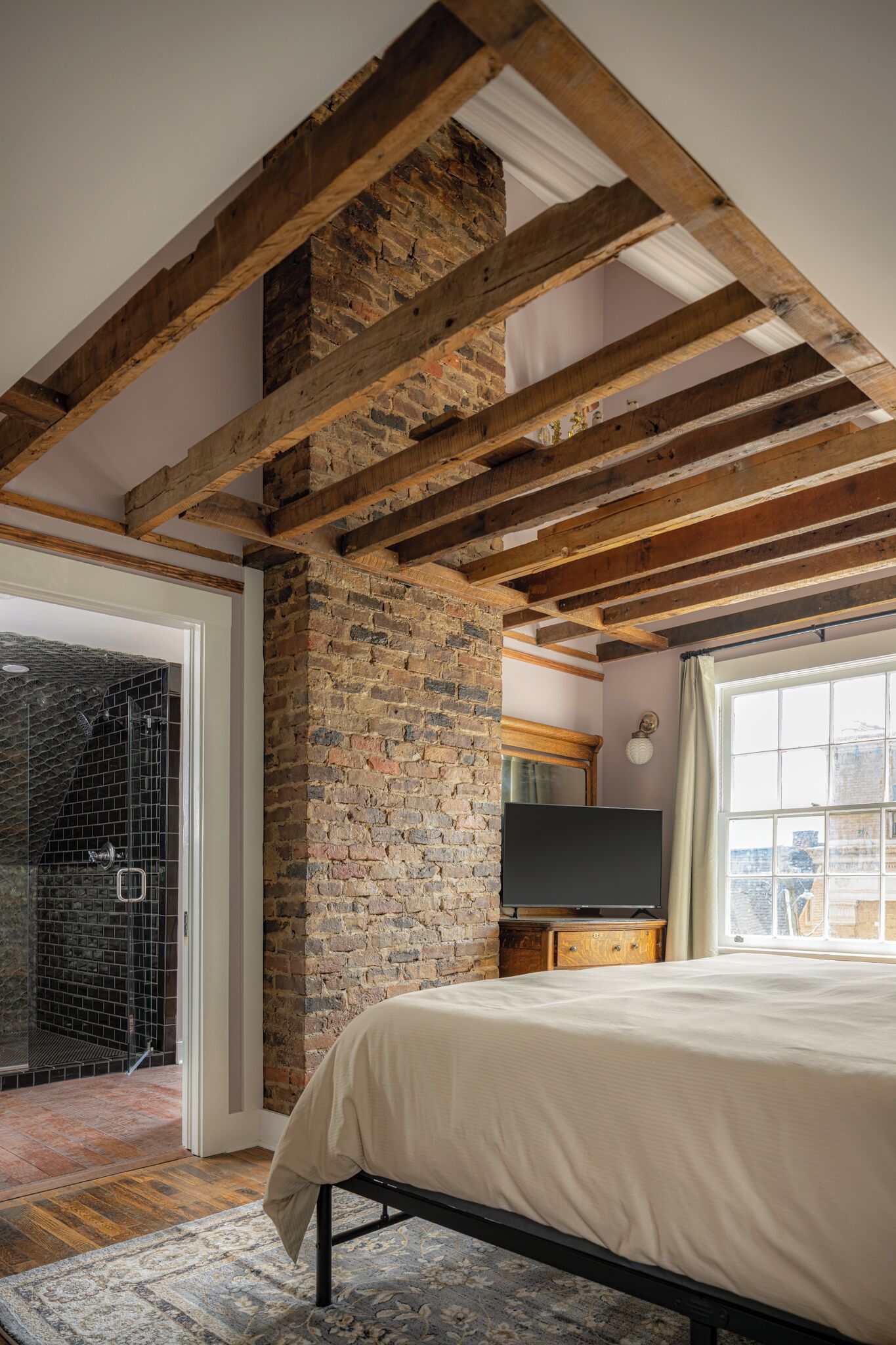 Un dormitorio con vigas de madera a la vista, una pared de ladrillo, una cama, un televisor y una ventana con vistas.