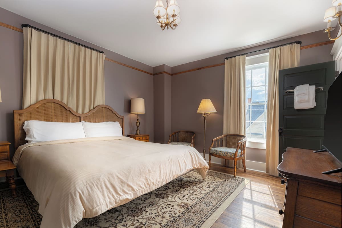 Ein Hotelzimmer mit einem großen Bett, zwei Nachttischen, einem Sessel, einem Schreibtisch, einer Lampe, einem Kronleuchter und Blick ins Freie durch ein mit Vorhängen drapiertes Fenster.