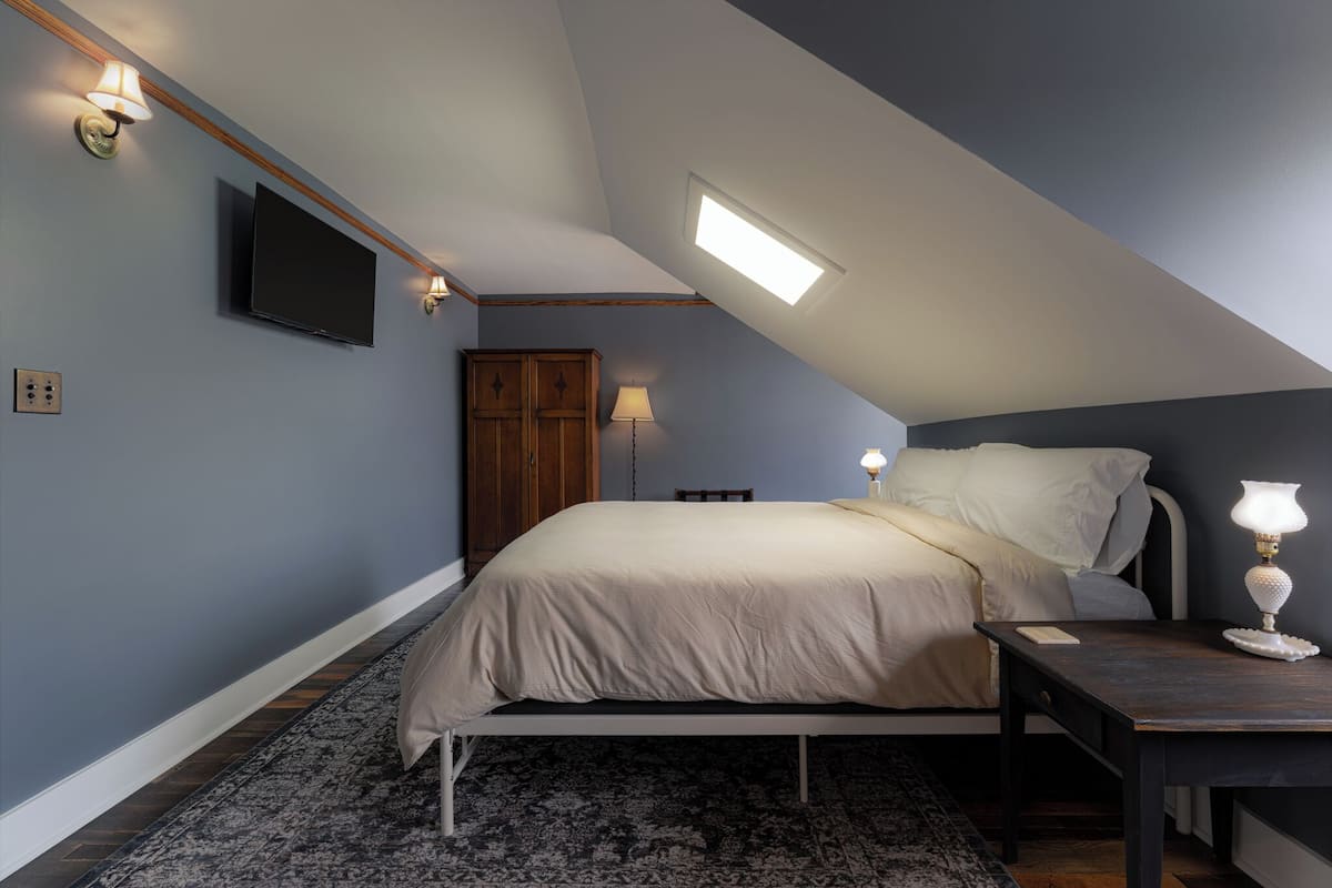 Ein Schlafzimmer mit einem Bett, einem Nachttisch, einer Lampe, einem an der Wand montierten Fernseher und einer Deckenleuchte.