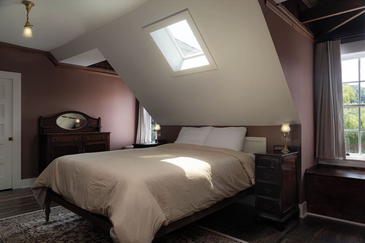 Ein Schlafzimmer mit schrägen Wänden, einem Oberlicht, einem Bett mit Kopfteil, einem Nachttisch, einer Kommode und einem Spiegel.
