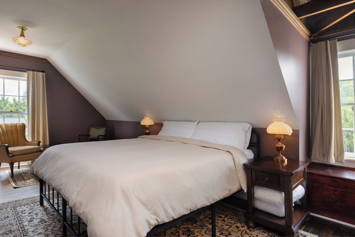 Ein Schlafzimmer mit einem großen Bett, zwei Nachttischlampen, einem Sessel und Blick ins Freie durch ein Fenster.