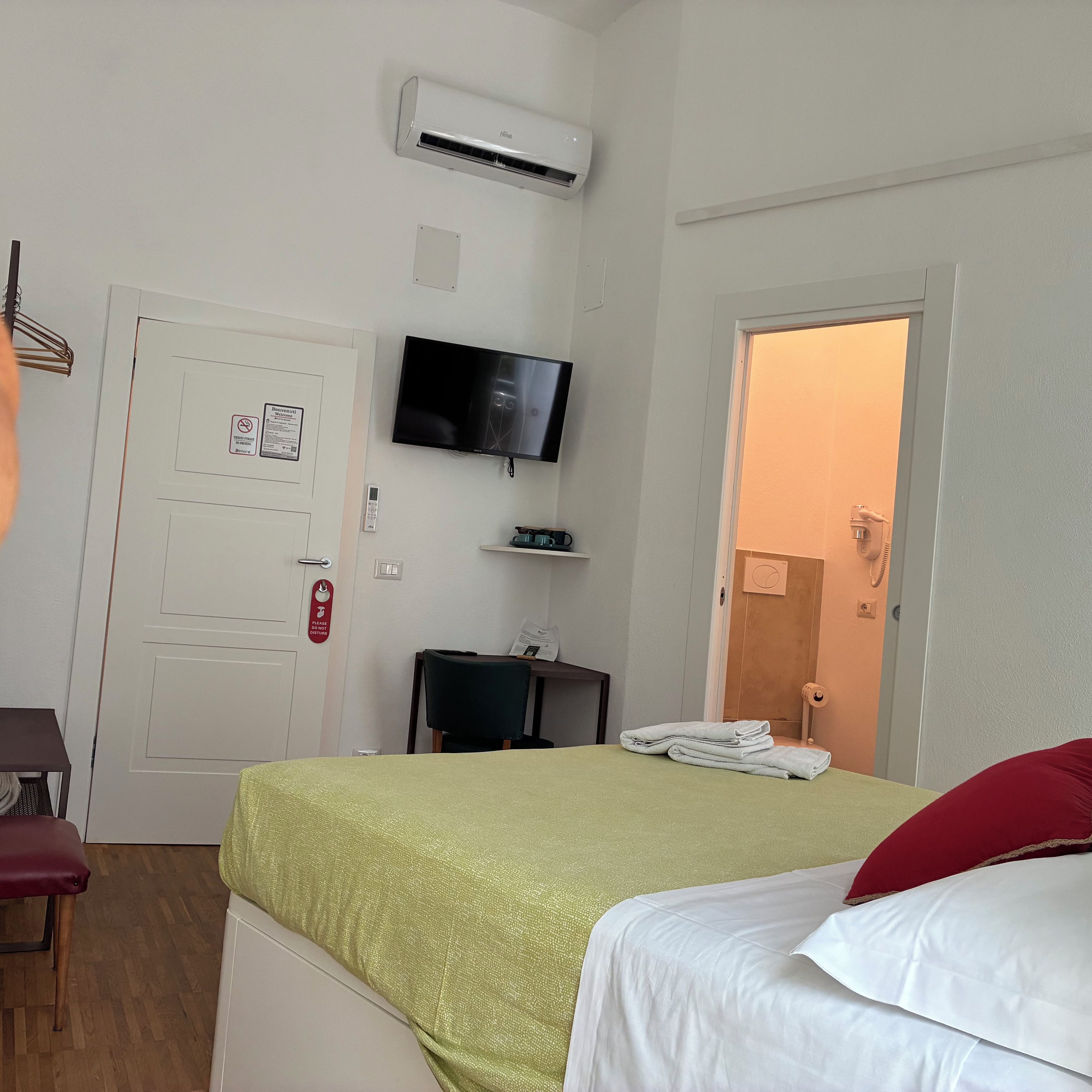 A hotel room with a bed, a television, an air conditioning unit, a desk, and a door leading to another room.