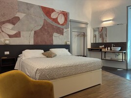 Un dormitorio moderno con una cama grande, una mesita de noche, una silla, un baño con lavamanos y espejo, y una pared con arte abstracto.