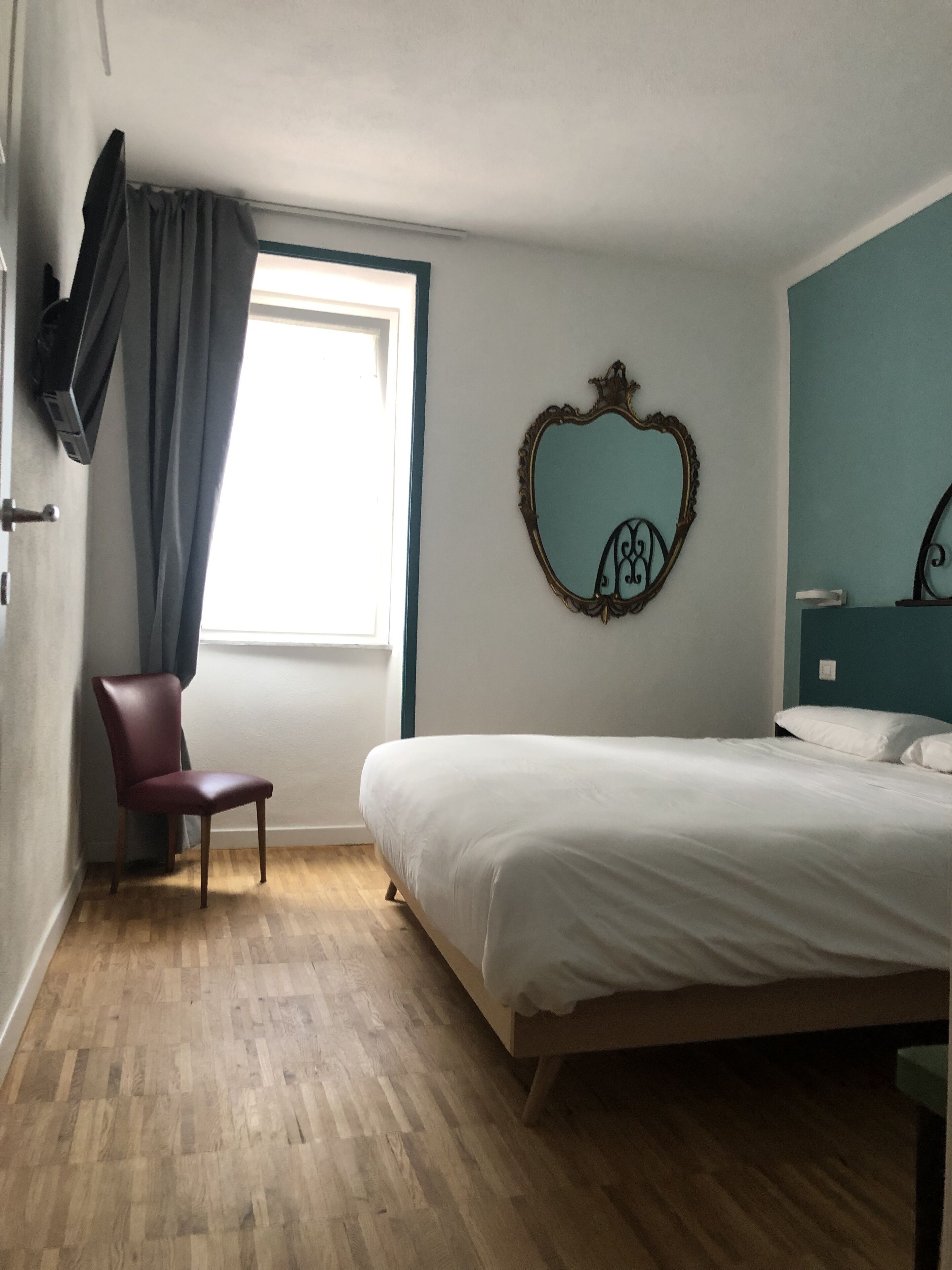 A bedroom with a bed, a chair, a window with curtains, and a wall-mounted mirror.