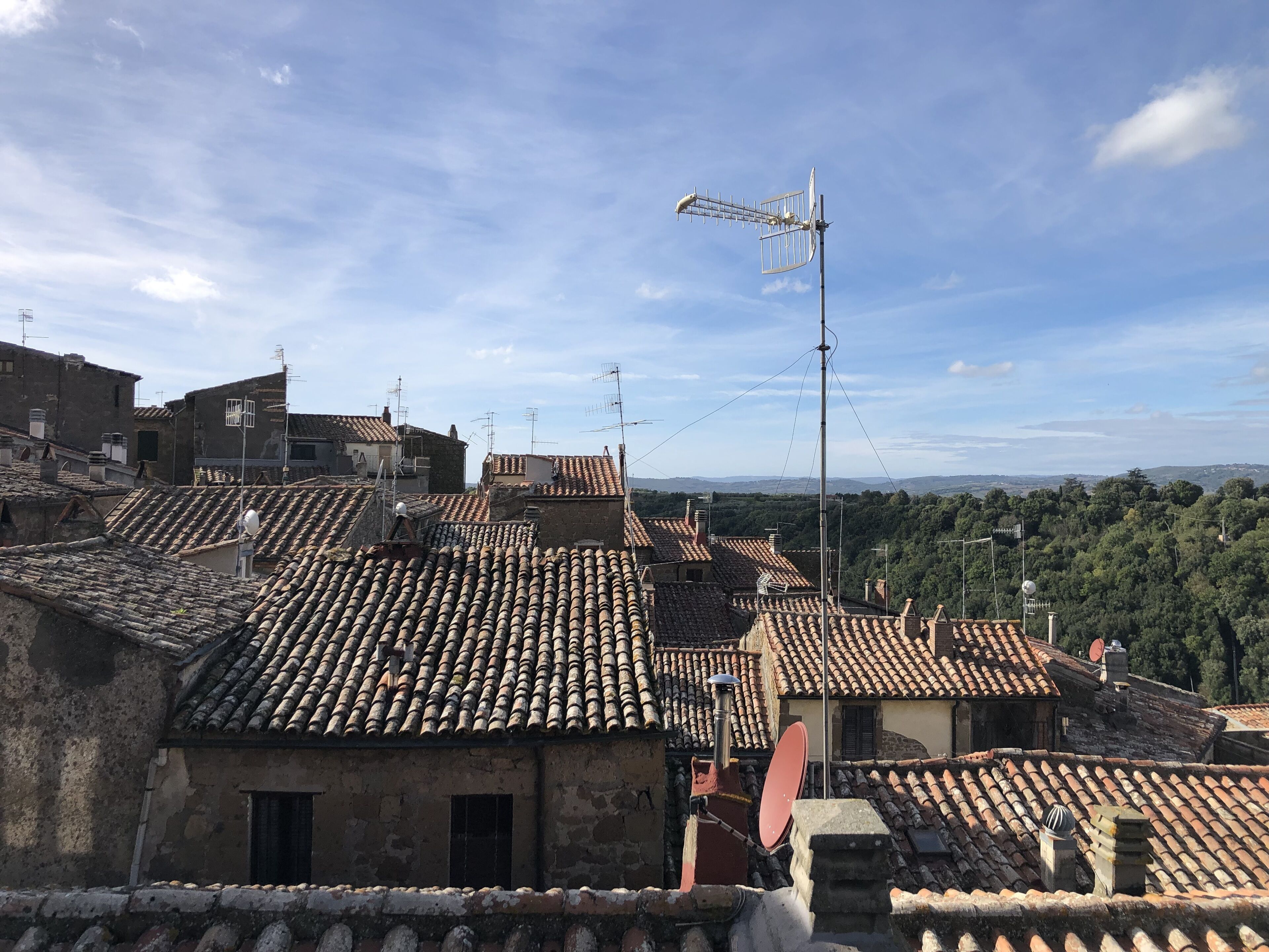 A view of terracotta-tiled rooftops with satellite dishes, surrounded by buildings with traditional architecture and a clear sky.