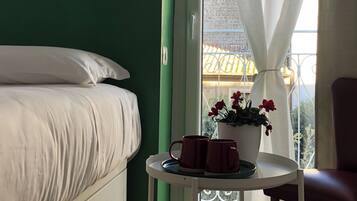 Ein Schlafzimmer mit einem Bett, einem kleinen Tisch mit zwei Tassen, einer Topfpflanze und Blick auf einen Balkon mit Geländer.