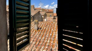 A view through open shutters of a rooftop with terracotta tiles, stone buildings, and a clear blue sky. - DIMOR'A'   ROOMS (Pitigliano)
