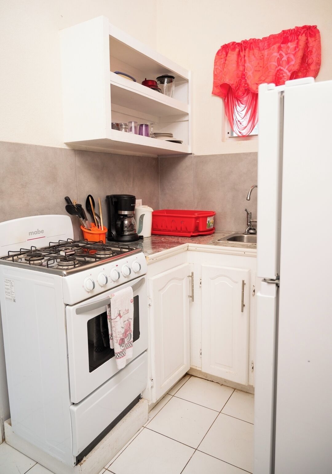 Een keuken met een kookplaat, oven, spoelbak en een koelkast. Er zijn kastjes en planken boven het aanrecht.
