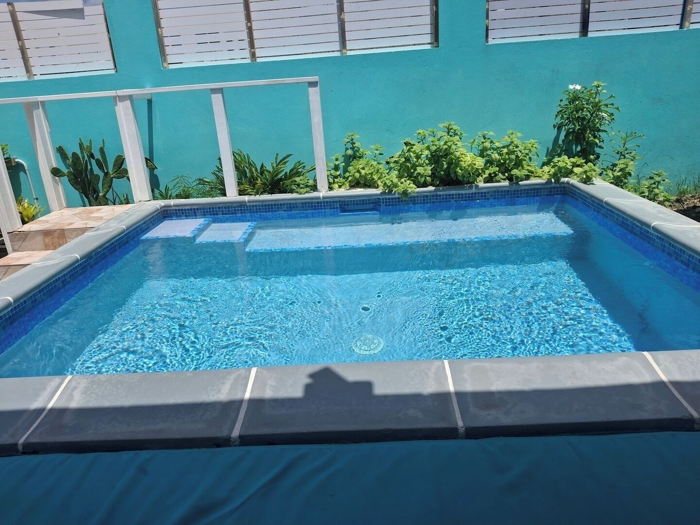 Een rechthoekig zwembad met helderblauw water, omringd door een betegeld zonnedek en een witte hek.