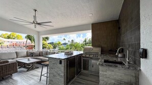 Cocina exterior con una parrilla, taburetes de bar y una zona de estar con mesa y sillas.