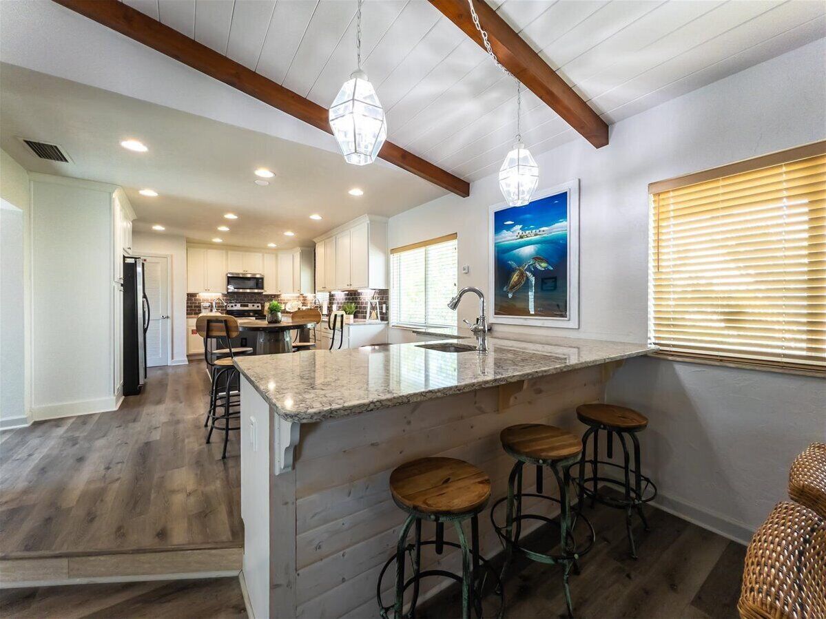 A spacious kitchen with a central island, wooden bar stools, pendant lighting, and a large window with blinds.