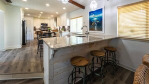 Cocina amplia con una isla central, taburetes de madera, iluminación colgante y un ventanal con persianas.