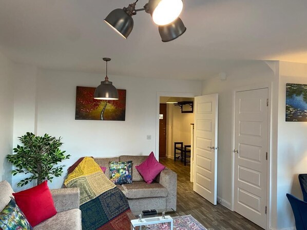 Sala de estar com sofá, mesa de centro e planta. Há uma porta que dá para outro cômodo.