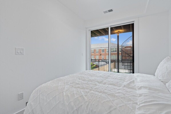 A bedroom with a large bed, white walls, and a view of a building through a sliding door. - Charming DC Retreat | Perfect for Sightseeing & Corporate Stays (Washington)