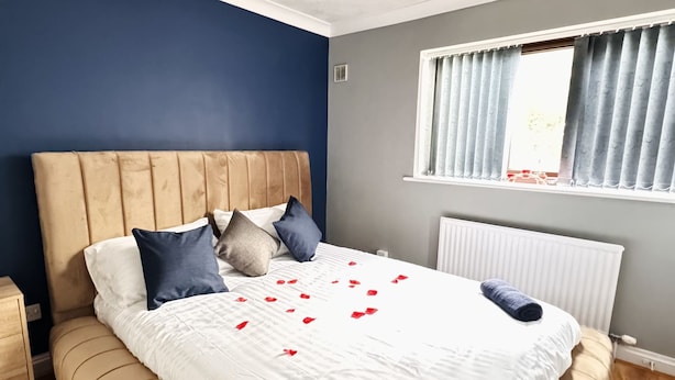 Een net opgemaakt bed met een witte dekbedovertrek, meerdere kussens en rozenblaadjes verspreid over het bed.