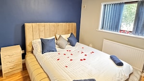 Een net opgemaakt bed met witte beddengoed en blauwe kussens, een houten nachtkastje en een raam met gordijnen.