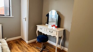 Een kamer met een houten vloer, een wit bureau met lades, een blauwe kruk, een klok aan de muur en een raam met gordijnen.