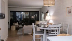 En matplats med ett runt bord och vita stolar, en hängande lampa och utsikt in i köket genom en glasdörr.