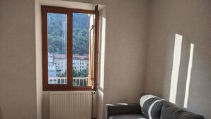 Une pièce avec une fenêtre donnant sur un paysage urbain, un radiateur blanc, un canapé gris et une table blanche.