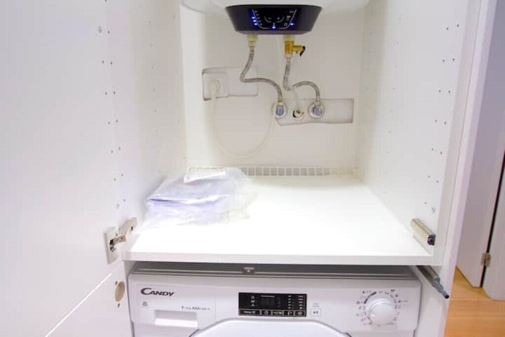 A compact, white laundry room with a Candy brand washing machine, a control panel, and a ceiling-mounted water heater.
