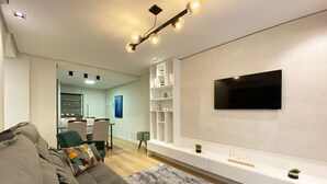 A modern living room with a sofa, a TV mounted on the wall, a dining area in the background, and a white console table with shelves.